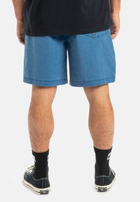 Pantaloni scurți din bumbac albastru, cu croială dreaptă, tiv rulat și un buzunar pe spate cu un singur nasture. Purtați cu șosete negre și pantofi.