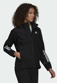 Czarna kurtka zapinana na zamek, wykonana z lekkiego materiału. Posiada trzy białe paski na rękawach oraz logo Adidas na klatce piersiowej.