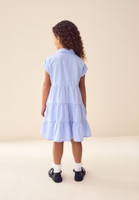 Mėlynos ir baltos spalvų languota suknelė su sluoksniuota sijono dalimi, trumpomis rankovėmis ir apykakle. Pora juodų batų ir baltos kulkšnies kojinės.