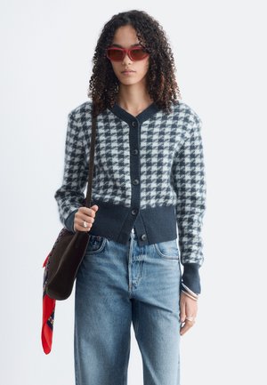 Femme aux cheveux bouclés portant des lunettes de soleil rouges, un cardigan à carreaux noir et blanc, un jean bleu et un sac foncé avec une écharpe rouge.