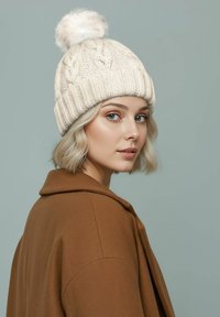 Cream knit beanie with a furry pom-pom on top, featuring a ribbed texture. The model wears a brown coat with a notched collar.