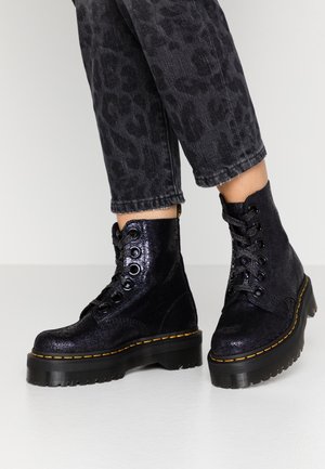 Zwarte glanzende veterplatformlaarzen gedragen met korte donkere luipaardprint jeans op een effen lichte achtergrond.