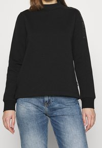 En kvinna bär en svart långärmad sweatshirt tillsammans med blå jeans, med en hand vilande på höfterna, vilket framhäver en avslappnad stil.