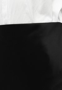 Η μαύρη φούστα έχει λεία υφή και συνδυάζεται με ένα λευκό πουκάμισο, δημιουργώντας έντονη αντίθεση στο χρώμα και έναν απλό, στρωματοποιημένο σχεδιασμό.