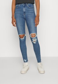 Vaqueros skinny azules con desgastes en las rodillas, de cintura alta y un diseño clásico de cinco bolsillos, combinados con zapatillas blancas.