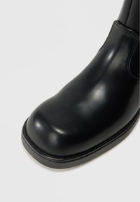 Botín de cuero negro hasta el tobillo con punta redondeada, textura suave, brillo sutil y diseño minimalista. Cuenta con una suela de perfil bajo.