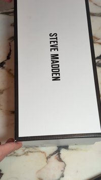 Eine Hand hält eine schwarz-weiße Steve Madden Schuhbox auf einer Marmoroberfläche.