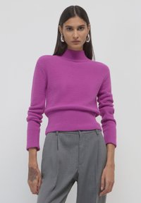 Maglione fucsia a maglia con colletto alto e polsini a costine, abbinato a pantaloni grigi chiari su misura. Morbida texture e design aderente.