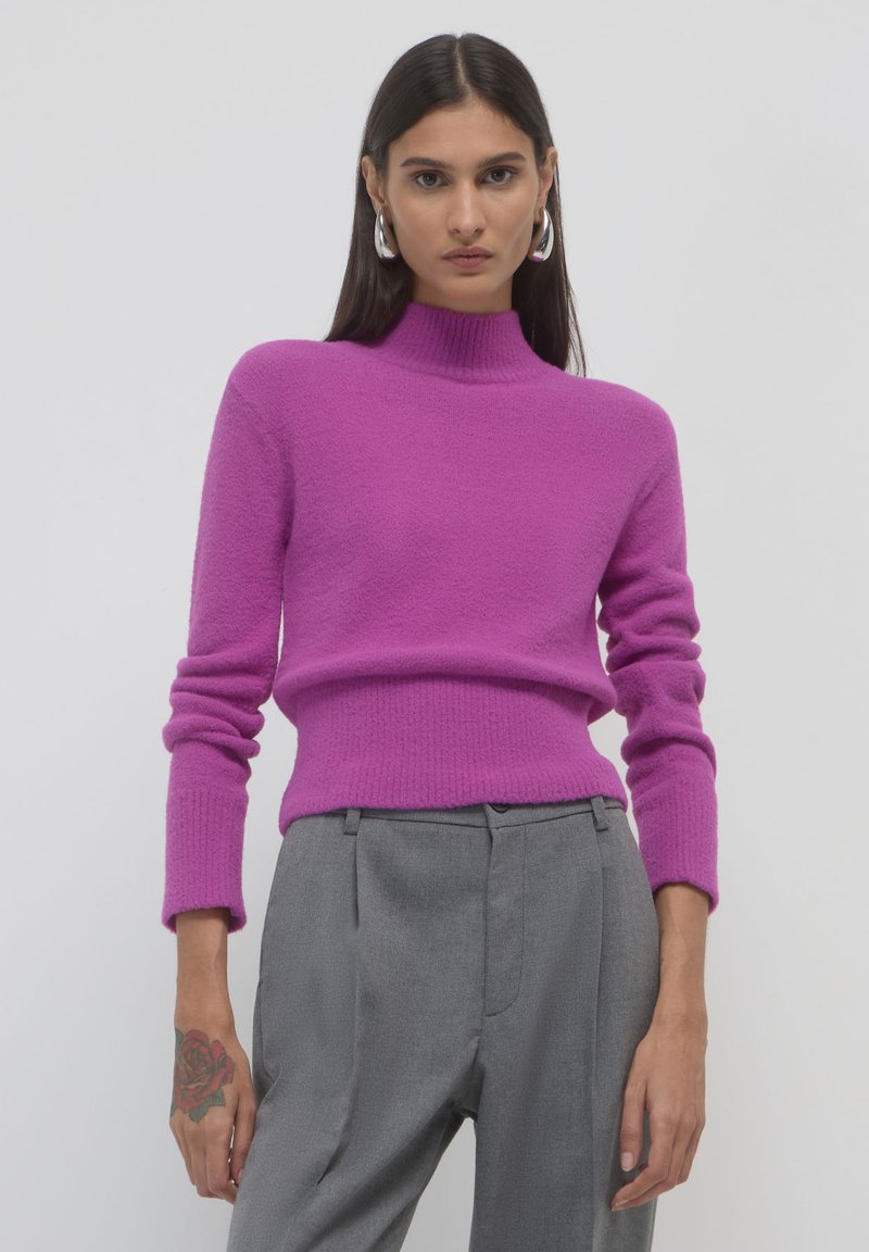 Maglione fucsia a maglia con colletto alto e polsini a costine, abbinato a pantaloni grigi chiari su misura. Morbida texture e design aderente.