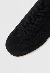 Μαύρο sneaker από σουέντ με στρογγυλεμένη μύτη, χοντρά μαύρα κορδόνια και λεπτομερείς ραφές στο επάνω μέρος πάνω σε ανοιχτό φόντο.