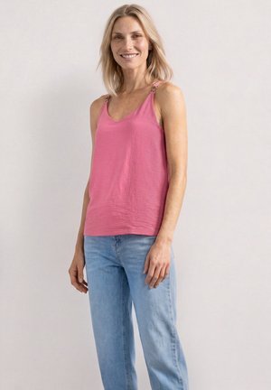Smilende kvinde med blondt hår iført en lyserød ærmeløs top og lyseblå jeans, stående foran en ensfarvet lys baggrund.