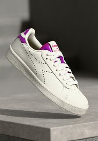 Hvit lær sneakers med lilla detaljer, rund tå, flat såle og perforerte detaljer. Har snøring og merke på tungen.