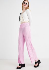 Rozā, plisētas, platas bikses, izgatavotas no viegla auduma, ar augstu elastīgu jostu un maziem vertikāliem svītrām. Valkātas melnas platformas sandales.