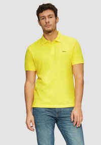 Gelbes Polohemd aus Baumwolle mit klassischem Kragen und kurzen Ärmeln. Verfügt über ein kleines Logo auf der linken Brust. Getragen mit blauen Jeans.