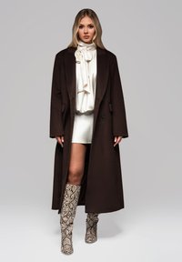 Ruskea villasekoite takki, jossa on pitkät hihat ja nappisulku, yhdistetty valkoiseen korkeakauluksiseen puseroon ja kuvioituihin polven ylle ulottuviin käärmeennahkakuosisin bootseihin.