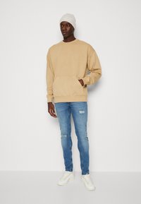 Beige sweatshirt met een voorzak, gecombineerd met lichtblauwe ripped jeans en witte sneakers. Het model draagt een lichtgrijze gebreide beanie.