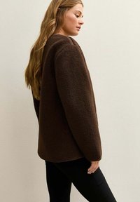 Chaqueta de forro polar marrón con un diseño cuadrado, textura suave y hombros caídos. Presenta una silueta simple y limpia sin adornos.