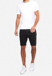 Homme portant un t-shirt blanc uni, un short noir à cordon de serrage et des baskets blanches, debout devant un fond clair.