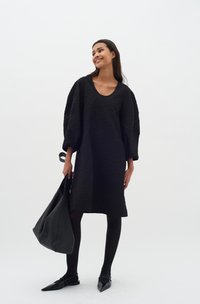 Schwarzes gestepptes Kleid mit weiten Ärmeln und Rundhalsausschnitt, kombiniert mit schwarzen Strumpfhosen und spitzen Ballerinas, dabei eine große schwarze Tasche haltend.