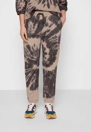 Person iført beige og mørkebrune tie-dye joggingbukser med elastiske manchetter og flerfarvede sneakers, stående mod en ensfarvet baggrund.