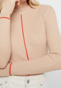 Beige geribbelde långarmige top met een hoge hals. Heeft een centrale rode lijn en rode accenten op de manchetten. Zachte textuur.