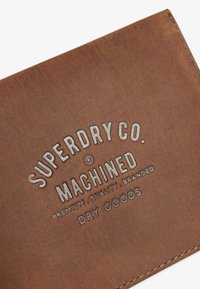 Cartera de cuero marrón con texto plateado en relieve, presenta el nombre de la marca y un eslogan, textura suave y costuras ordenadas en los bordes.