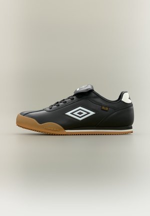Fekete bőrből készült alacsony szárú sportcipő gumitalppal, fehér Umbro logó az oldalon, fekete fűzők és a nyelv fölött egy hajtóka semleges háttér előtt.