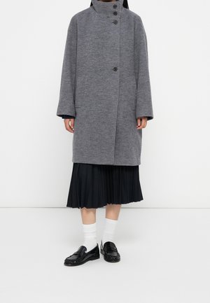 Person iført en lang grå knappet frakke over en sort plisseret nederdel, hvide sokker og sorte læder loafers mod en ensfarvet baggrund.