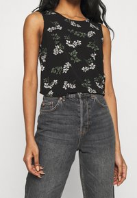 Zwarte mouwloze top met wit bloemmotief, gecombineerd met hoge donkergrijze jeans. Glad materiaal, ontspannen pasvorm, minimale details.