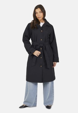 Donna con capelli lunghi e scuri che indossa un trench nero con cintura, jeans azzurri a gamba larga e scarpe nere con punta, su sfondo bianco.