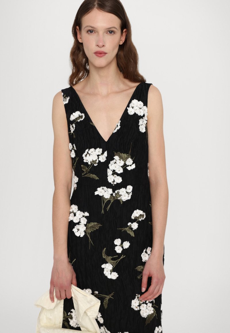 Robe noire à décolleté en V profond, ornée de motifs floraux blancs et de feuilles vertes. Le mannequin tient un tissu crème dans une main.