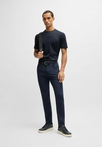 T-shirt girocollo navy abbinato a pantaloni slim-fit blu scuro, una cintura nera e sneakers con suola chiara. Modello con una borsa nera.