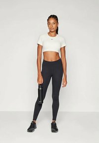 Witte crop top, zwarte high-waisted leggings en zwarte sneakers. Houdt een zwarte waterfles vast. Glad stof met een aansluitend ontwerp.