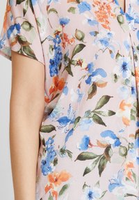 Lyserød bluse med blomsterdesign i blå, orange og grønne nuancer. Korte ærmer og struktureret stofdetalje.