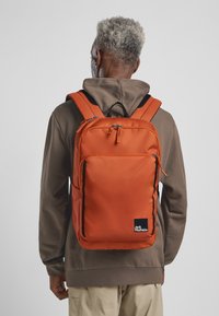 Oranje rugzak met meerdere vakken, zwarte accenten en logo. Gemaakt van duurzaam materiaal met gewatteerde banden en een gestructureerd oppervlak.