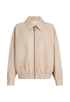 Frau geht nach vorne, trägt eine beige Jacke, cremefarbene weite Hosen, Sandalen und trägt eine große cremefarbene Tragetasche vor einem einfarbigen grauen Hintergrund.