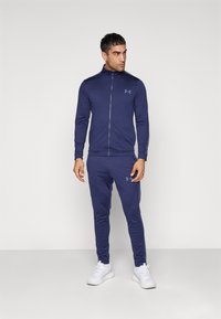 Tamnoplavi sportski trenirka napravljena od glatkog materijala, s jaknom na zip te uskim hlačama s diskretnim logotipom. Bijele tenisice upotpunjuju izgled.