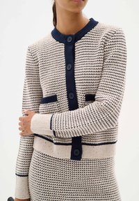 Knit cardigan in cream with navy trim, featuring a round neck, five buttons, and two front pockets. Textured surface with horizontal stripes.