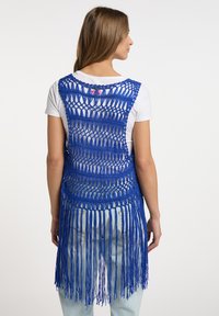 Blå makraméväst med fransdetaljer, med en öppen vävdesign. Bärs över en vit skjorta, vilket framhäver dess textur och hantverk.