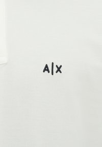 Baltas polo marškinėliai su apykakle, pagaminti iš tekstūruoto audinio; turi mažą juodą siuvinėtą "A|X" logotipą kairėje krūtinės pusėje.