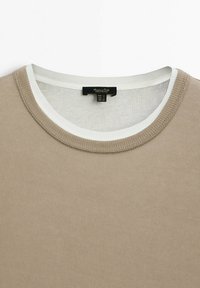 Pull beige à col rond superposé sur un t-shirt blanc à col rond, montrant des coutures détaillées et une étiquette de vêtement au niveau de l'encolure.