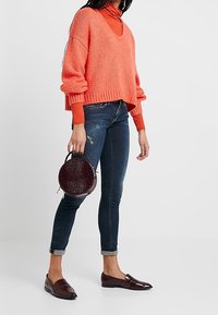 Orange stickad tröja med v-ringning, lagd över en orange polotröja. Mörkblå figursydda jeans, bruna läderloafers och en rund vinröd handväska.