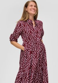 Bordeaux jurk met een geometrisch wit patroon, kraagontwerp, korte opgerolde mouwen en een tiered ruffle zoom. Bevat zijzakken.