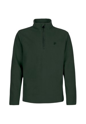 Junge Person trägt eine schwarze Mütze, dunkelgrünes Langarmshirt, leuchtend orange Hose und schwarze Stiefel vor einfarbigem Hintergrund.