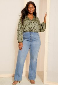 Grüne Bluse mit Polka-Dots, V-Ausschnitt und lockeren Ärmeln, kombiniert mit hoch taillierten hellblauen Jeans und beigen Riemchensandalen.
