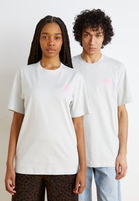 Ljust grå bomullströjor med korta ärmar, med en rosa Adidas-logotyp på bröstet, i kombination med avslappnade kläder, placerade mot en neutral bakgrund.