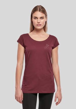 Jeune femme aux cheveux blonds et raides porte un t-shirt bordeaux à manches courtes avec un petit motif cerise sur le côté gauche de la poitrine.