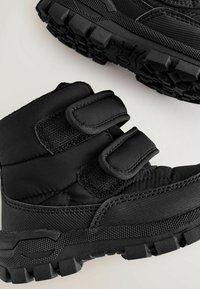 Sorte vinterstøvler med et polstret skaft, der har velcro-remme, en struktureret sål og gummiafsætninger for bedre fodfæste. Holdbart syntetisk materiale.