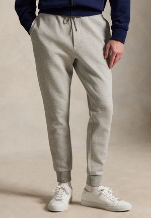 Mężczyzna ubrany w jasnoszare spodnie dresowe i białe trampki, stojący z jedną ręką w kieszeni na neutralnym tle.