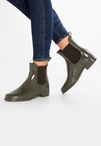 Persona vistiendo botas de lluvia brillantes de color verde oscuro hasta el tobillo y jeans ajustados azules, contra un fondo blanco liso.